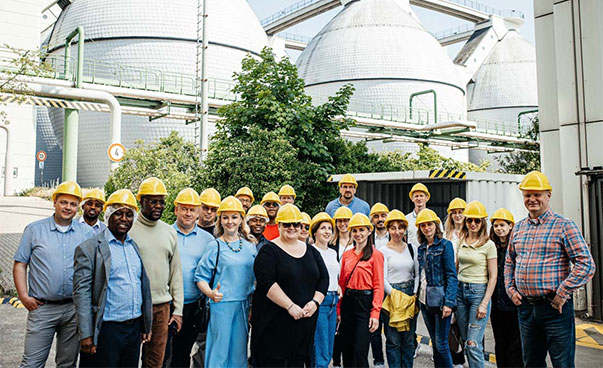 About two dozen people in yellow construction helmets stand in front of several large metal containers and smile at the camera.