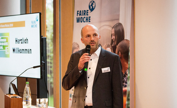 Ein Mann steht und redet in ein Mikrofon. Im Hintergrund ist die Aufschrift "Faire Woche" zu sehen. Foto: Forum Fairer Handel