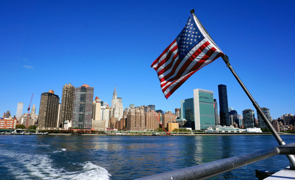 Blick von einem Boot mit einer US-amerikanischen Flagge auf die Skyline von New York, mit dem UN-Gebäude rechts.