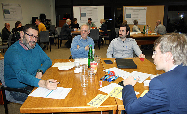 Eine Arbeitsgruppe sitzt um einen Tisch und diskutiert. Im Hintergrund sind weitere Arbeitsgruppen zu sehen.