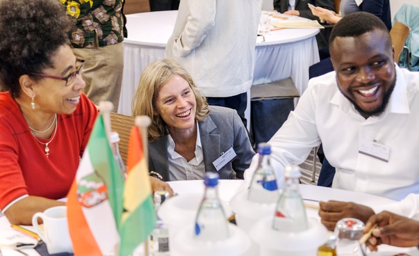 Zwei Frauen und ein Man sitzen an einem Tisch mit Konferenzgetränken und lachen. Im Vordergrund sind unscharf die Flaggen von Ghana und NRW zu sehen.