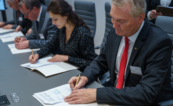 Drei Männer und eine Frau sitzen an einem langen Tisch und unterzeichnen Urkunden.