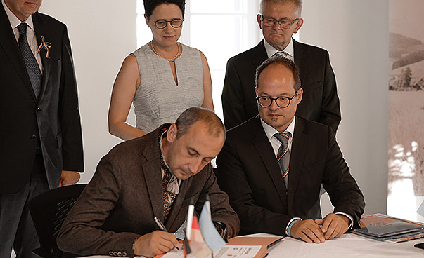 Mykhailo Tshuliak, Bürgermeister von Trostjanez und Martin Aßmuth, Bürgermeister von Hofstetten sitzen nebeneinander und unterzeichnen einen Vertrag. Drei Menschen stehen im Hintergrund und schauen zu.