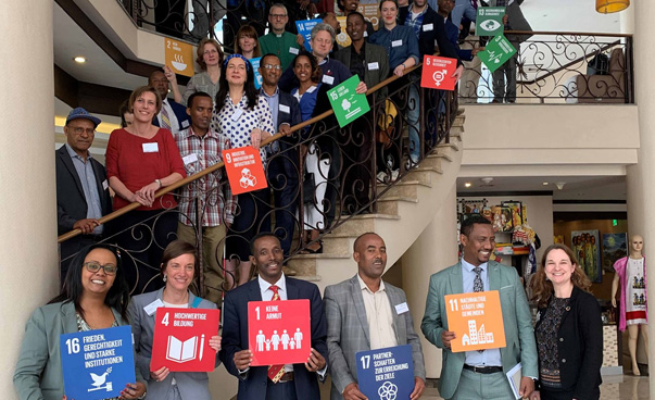 Approximately 30 people can be seen. Some of them are standing on a staircase, holding up banners with the symbols of the global sustainability goals.