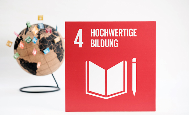 Das rote SDG-Logo Nummer vier "Hochwertige Bildung" mit einem Buch und einem Stift ist vor einem mit Fähnchen besteckten, unscharfen Globus zu sehen.