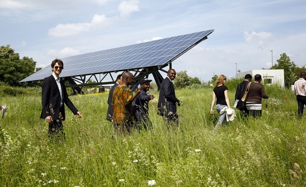 Mehrere Personen laufen durch hohes Gras, einige lächeln in die Kamera. Im Hintergrund steht schräg auf dem Boden ein sehr großes Solarmodul.
