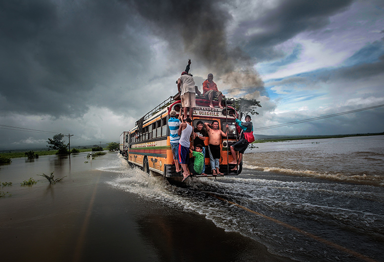 Un bus, sur lequel plusieurs personnes s'accrochent à l'extérieur à l'arrière, roule sur une route inondée ; dans le ciel, on peut voir des nuages très sombres.