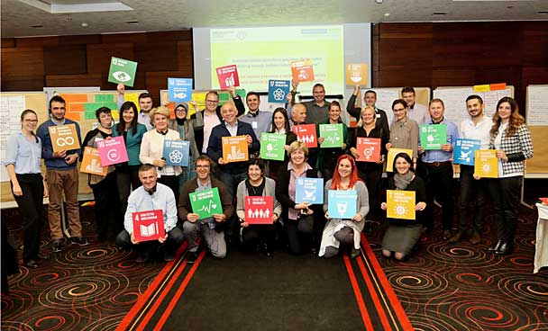 This group photo shows participants at the network meeting of 'Municipal Partnerships for Sustainability' in Sarajevo. Participants are standing in the conference room in front of partitions, holding up boards showing the 17 SDGs.