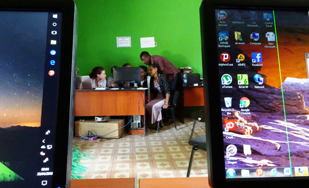 Durch die Lücke zwischen zwei Tablets blickt man auf eine Gruppe von Männern und Frauen, die gemeinsam auf einen Computer-Monitor schauen.