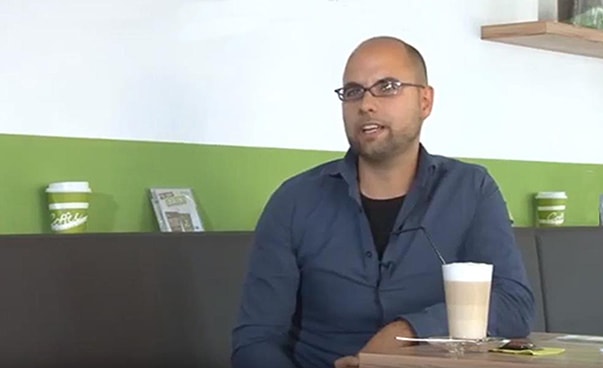Ein Mann spricht zu einem Interviewpartner; er sitzt in einem Café, vor ihm steht ein Glas mit Milchkaffee.