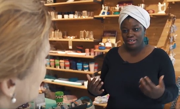 Eine Frau mit weißem Turban steht in einem Ladenlokal vor einem Regal mit Lebensmitteln; sie spricht mit einer Frau, die nur im Halbprofil zu erkennen ist.