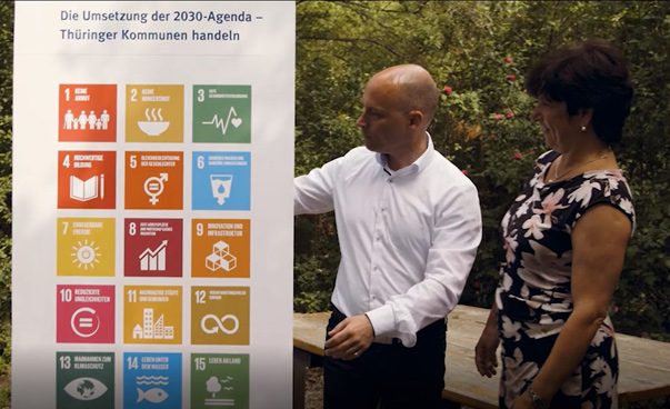 Ein Mann und eine Frau stehen lachend vor einem Plakat mit den Nachhaltigkeitssymbolen