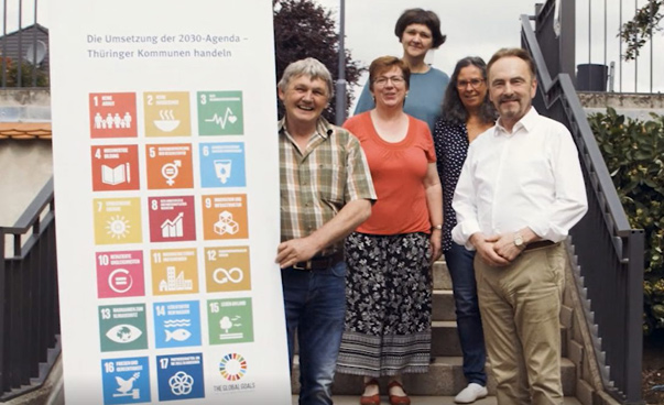 Mehrere Personen stehen lachend vor einem Plakat mit den Nachhaltigkeitssymbolen