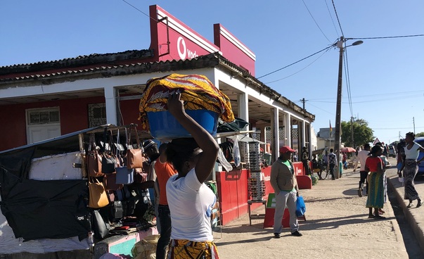 Straßenszene in Vilankulo. Im Vordergrund trägt eine Frau eine blaue Wanne mit Wäsche. Im Hintergrund ein Gebäude und mehrere andere Personen.