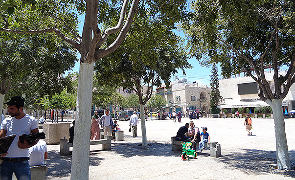 Auf dem Bild ist der Vorplatz der Bethlehem Peace Centers zu sehen, im Vordergrund sorgen vereinzelte Laubbäume für eine angenehme Begrünung des Platzes. Unter ihnen befinden sich Sitzbänke, auf denen sich Menschen tummeln. Im Hintergrund ist das zum Frie
