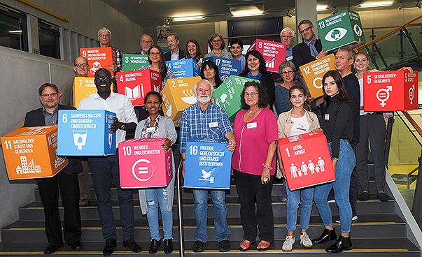 Menschen stehen zu einer Gruppe zusammen und halten Schilder mit den 17 Zielen für Nachhaltige Entwicklung hoch.