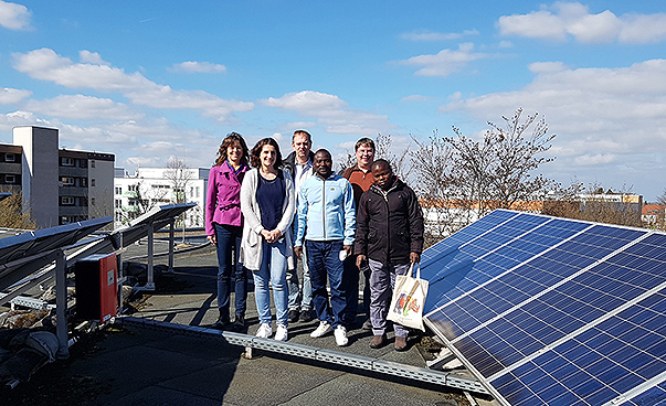 Sechs Menschen stehen auf dem Flachdach eines Hauses. Neben Ihnen befindet sich eine Solaranlage.