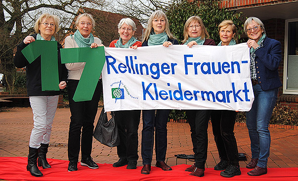 Sieben Frauen halten ein Schild mit der Beschriftung "17. Rellinger Frauen-Kleidermarkt" hoch