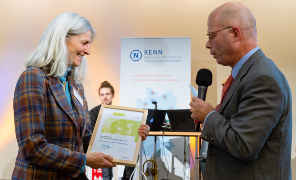 Ein Mann mit Mikrofon überreicht eine Urkunde an Elke Bröcker aus dem Rhein-Pfalz-Kreis. Foto: RENN.west