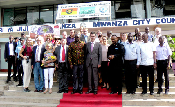 Eine große Gruppe steht auf einer Treppe und zum teil auf einem roten Teppich. Im Hintergrund an einem Gebäude ist abgeschnitten der Schriftzug Mwanza City zu sehen.
