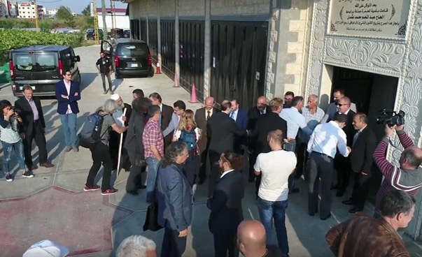 Videostandbild: Menschen stehen in Gruppen vor einem Gebäude zusammen