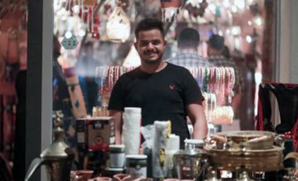 Video still: A man at a market stall smiles at the camera.
