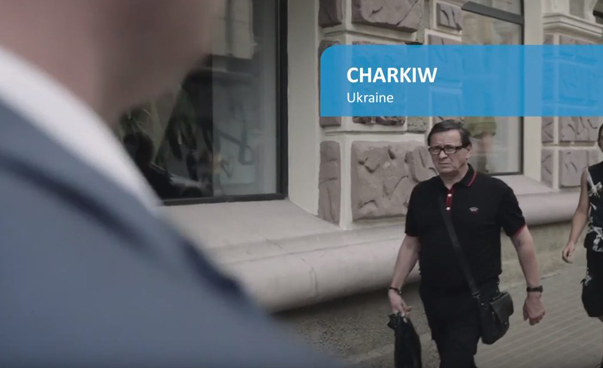 Ein Mann geht an einer Hauswand entlang auf den Betrachter zu. Links im Vordergrund ist verschwommen der Rücken eines Mannes zu sehen. Rechts oben zeigt ein blaues Banner den Namen Charkiw.