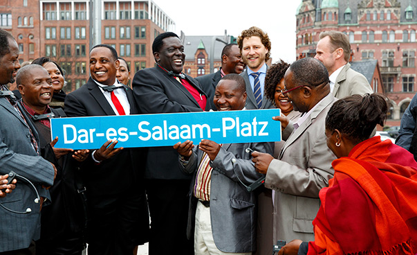 Eine Gruppe gutgelaunter Menschen hebt das Straßenschild Dar-es-Salaam-Platz in die Höhe.