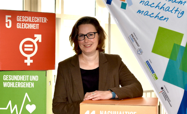 Porträt von Sonja Helmich neben bunten SDG-Würfeln und einer Dropflag der Stadt Rheinberg.
