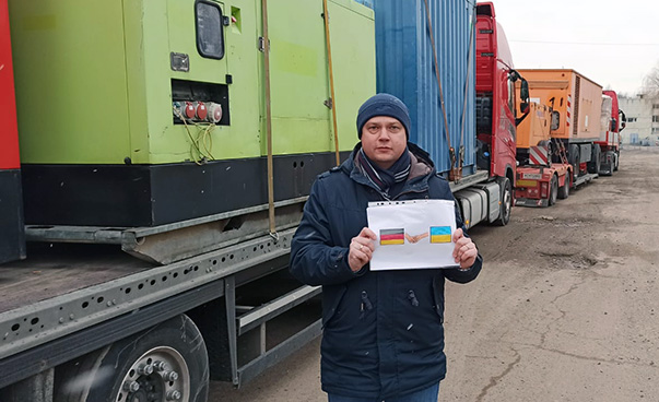 Volodymyr Bilynskyy stands in front of some trucks and holds a sign with a German and a Ukrainian flag into the camera.