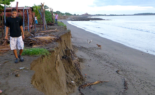 Blick auf einen abgebrochenen Küstenabschnitt; eine Person geht knapp an der Abbruchkante entlang; im Hintergrund ist eine Hütte zu sehen.