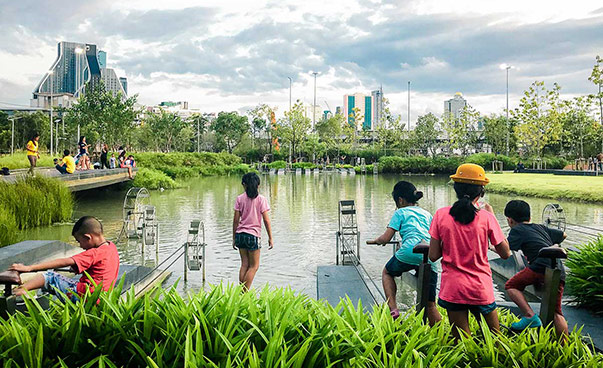 Kinder spielen in einem Park in Bangkok vor einer Wasserfläche; im Hintergrund sind Hochhäuser zu sehen.