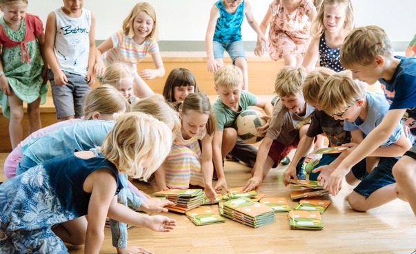 Rund 15 Mädchen und Jungen im Grundschulalter beugen sich über einen Stapel Hefte.