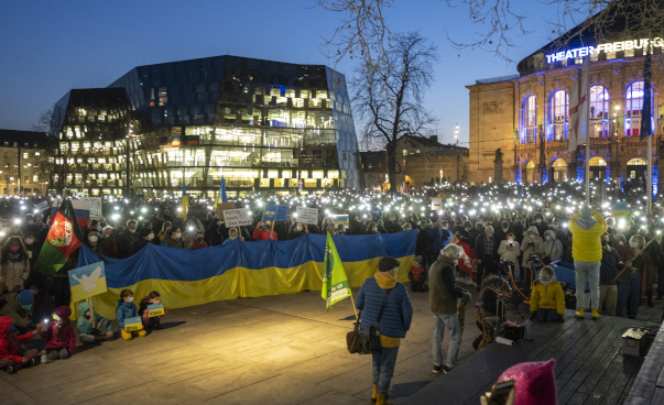 A group of people in semi-darkness, with lights, Ukrainian flags and banners. Behind them are two illuminated buildings, one is the theatre in Freiburg.