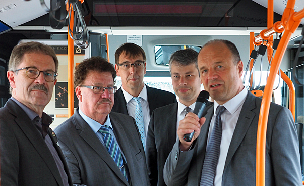 Five men stand side by side in a bus, one of them talking into a microphone.