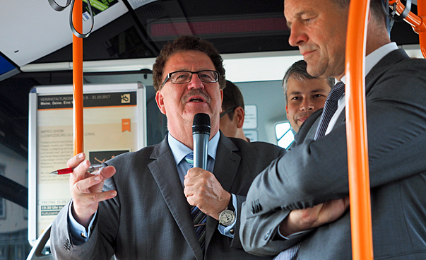 A man speaks into a microphone, he is with other people in a bus.
