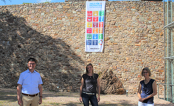 Drei Menschen stehen vor einer Mauer an der eine Fahne mit den 17 Zielen für Nachhaltige Entwicklung aufgehängt wurde.