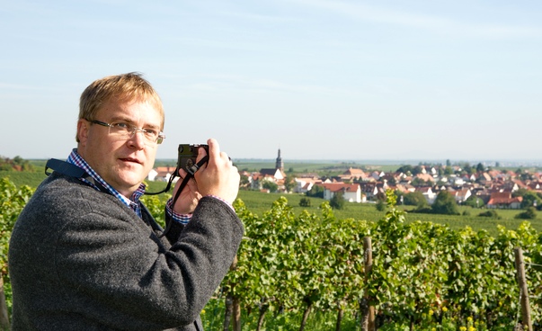 Der Kallstadter Bürgermeister Thomas Jaworek steht mit einem Fotoapparat in einem Weinberg. Im Hintergrund ist ein Dorf zu sehen.