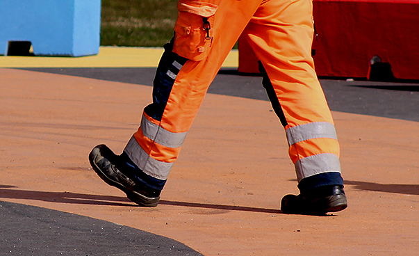 Die Arbeitsschuhe und das orangene Beinkleid eines Bauarbeiters sind zu sehen.