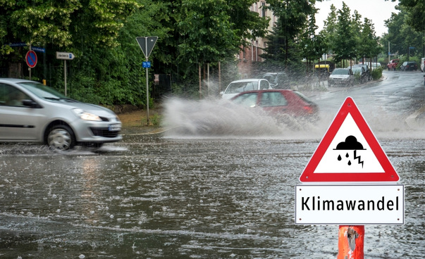 Zwei Autos fahren über eine überflutete Straße. Im Vordergrund warnt ein Schild vor der Klimakrise.
