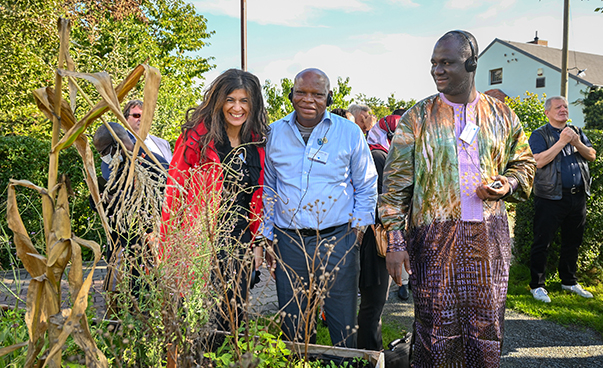 Teilnehmende der Konferenz erkunden eine Kleingartenanlage. Im Zentrum des Bildes stehen eine Frau und zwei Männer vor einem Gemüsebeet und lächeln in Richtung Kamera. Hinter ihnen stehen weitere Personen, die durch die Anlage spazieren.