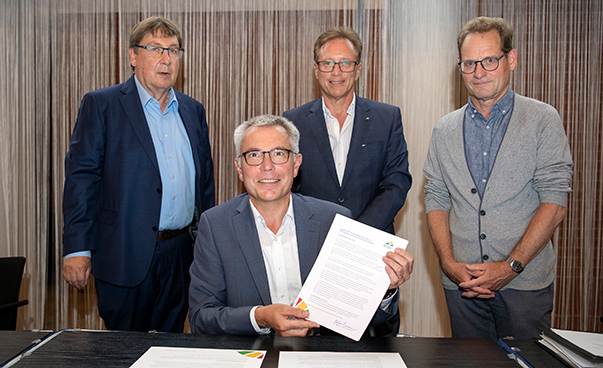 Stefan Dallinger, der Verbandsvorsitzende der Metropolregion Rhein-Neckar, sitzt an einem Tisch und hält die Musterresolution in die Höhe. Im Hintergrund stehen drei Männer