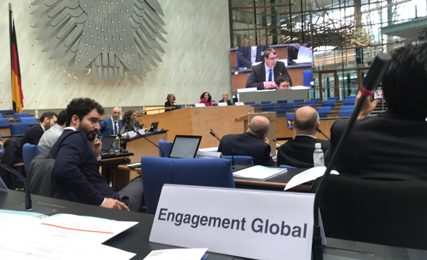 Im Plenarsaal des alten Bundestages vor dem Bundesadler spricht an Mann an einem Pult. Links ist ein weiterer Mann zu sehen, der sich umdreht. Vorne im Bild steht ein Schild mit der Aufschrift Engagement Global.