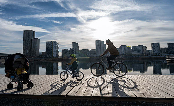 Ein Kind und ein Mann fahren auf einem Fahrrad über einen Holzsteg in der Wiener „Seestadt Aspern“; links steht ein Kinderwagen; im Hintergrund ist ein Gewässer und moderne, hohe Wohnhäuser zu sehen.