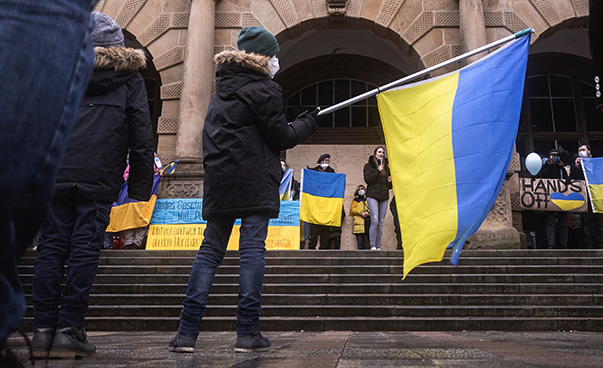 Vor einem historischen Gebäude mit einer Treppe im Vordergrund stehen mehrere Personen mit ukrainischen Flaggen.