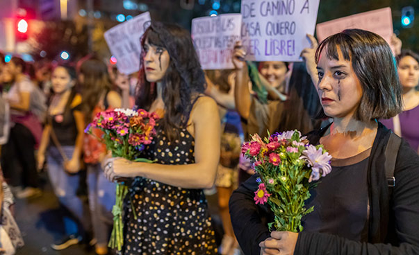 Dunkel gekleidete und geschminkte Frauen, mit Blumensträußen in der Hand, demonstrieren.