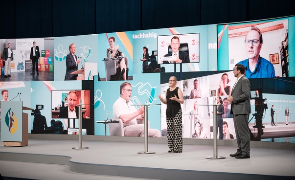 Bühne der 15. Bundeskonferenz für Kommunale Entwicklungspolitik mit der Moderatorin und dem Moderator. Im Hintergrund sind große Bilder der Konferenz auf einem Bildschirm zu sehen.