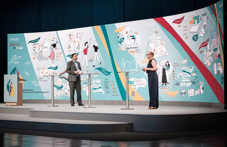 Der Moderator Alexander Thamm und die Moderatorin Julia Pfinder stehen auf der Bühne vor dem großen Screen, auf dem die Bundeskonferenz als Graphic Recording visuell dargestellt ist.