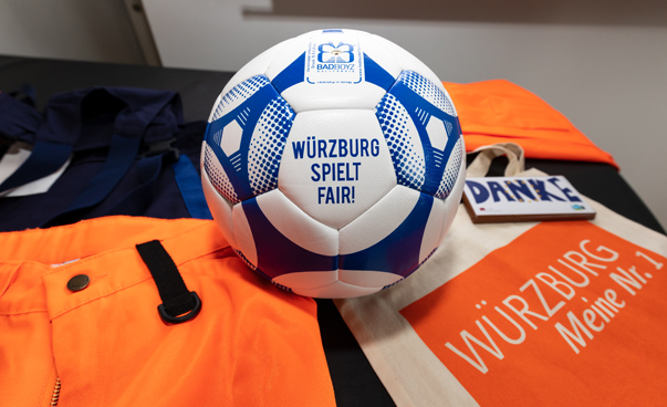 Im Zentrum des Bilder ist ein Lederfußball zu sehen mit der Aufschrift "Würzburg spielt fair". Ebenfalls ist der obere teil einer orange-farbenen Hose im Bild zu sehen.