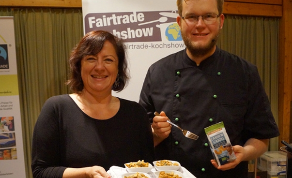 Eine Frau und ein Mann stehen mit Essen und Lebensmitteln vor einem Banner mit der Aufschrift Faire Kochshow. Foto: Brigitte Janosch